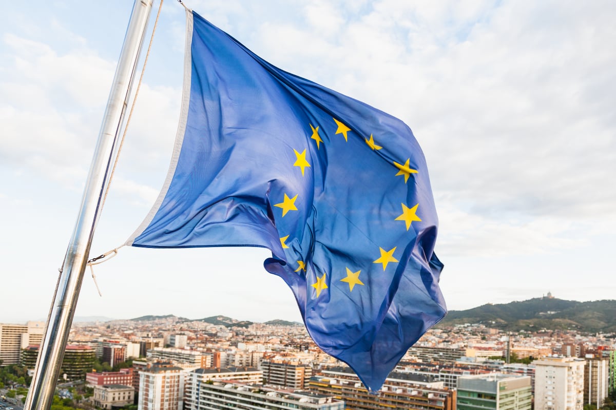 Vlag van de Europese Unie met gele sterren die wappert aan een vlaggenmast, met een stad en heuvels op de achtergrond.

