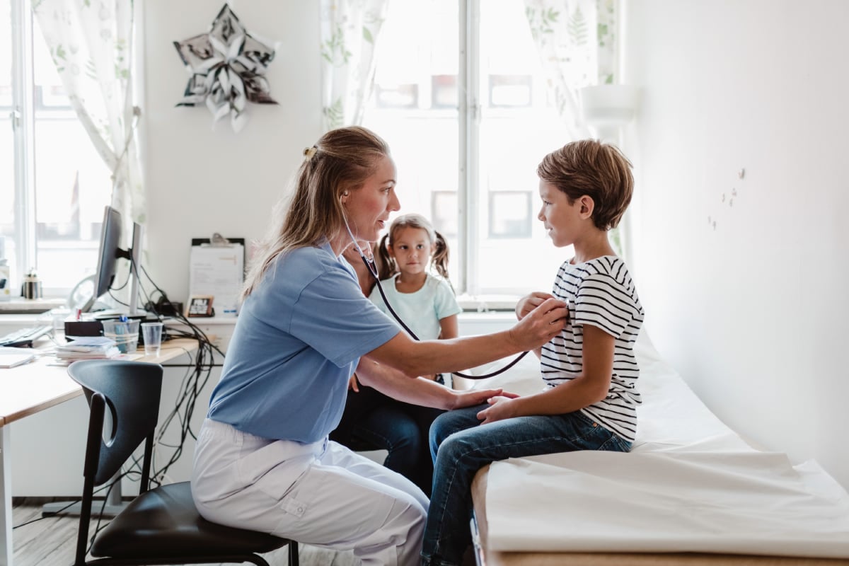 Healthcare professional using a stethoscope to examine a child, while another child stands in the background in a clinical room.


