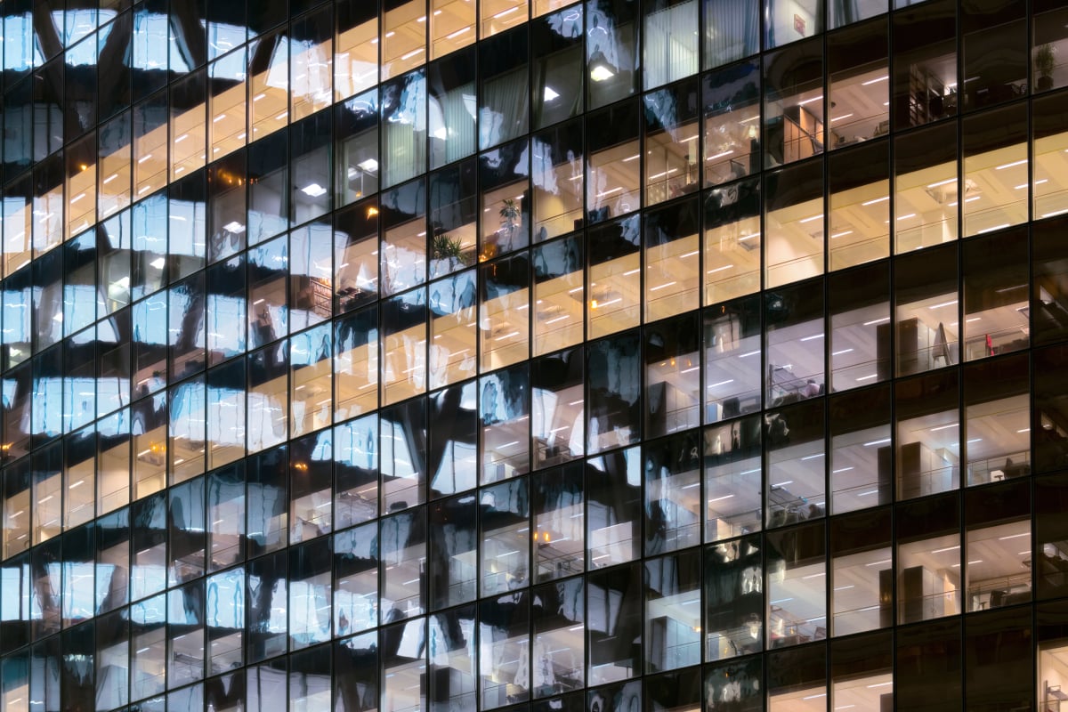 Glass facade of an office building with illuminated workspaces at night, illustrating corporate activity and financial markets
