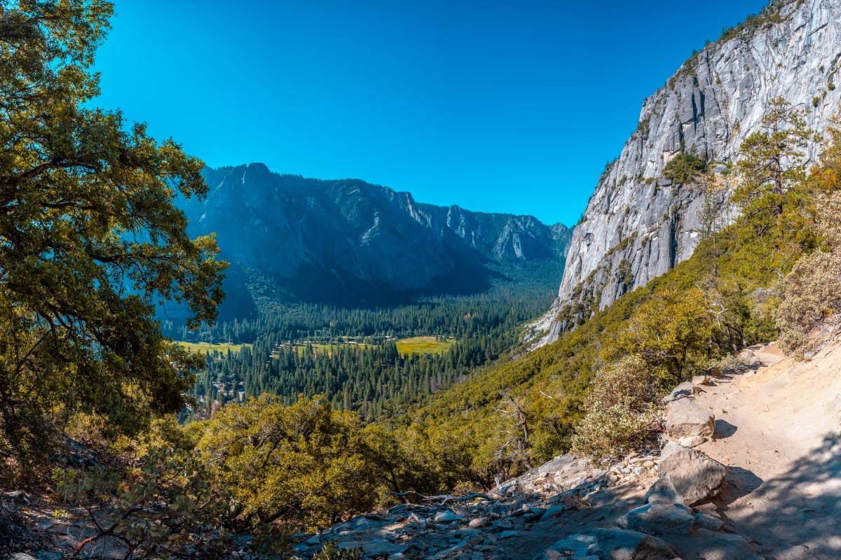 A scenic mountain valley viewed from a rocky hiking trail, with towering granite cliffs, dense evergreen forests, and a sunlit meadow below. The landscape is framed by trees under a clear blue sky.