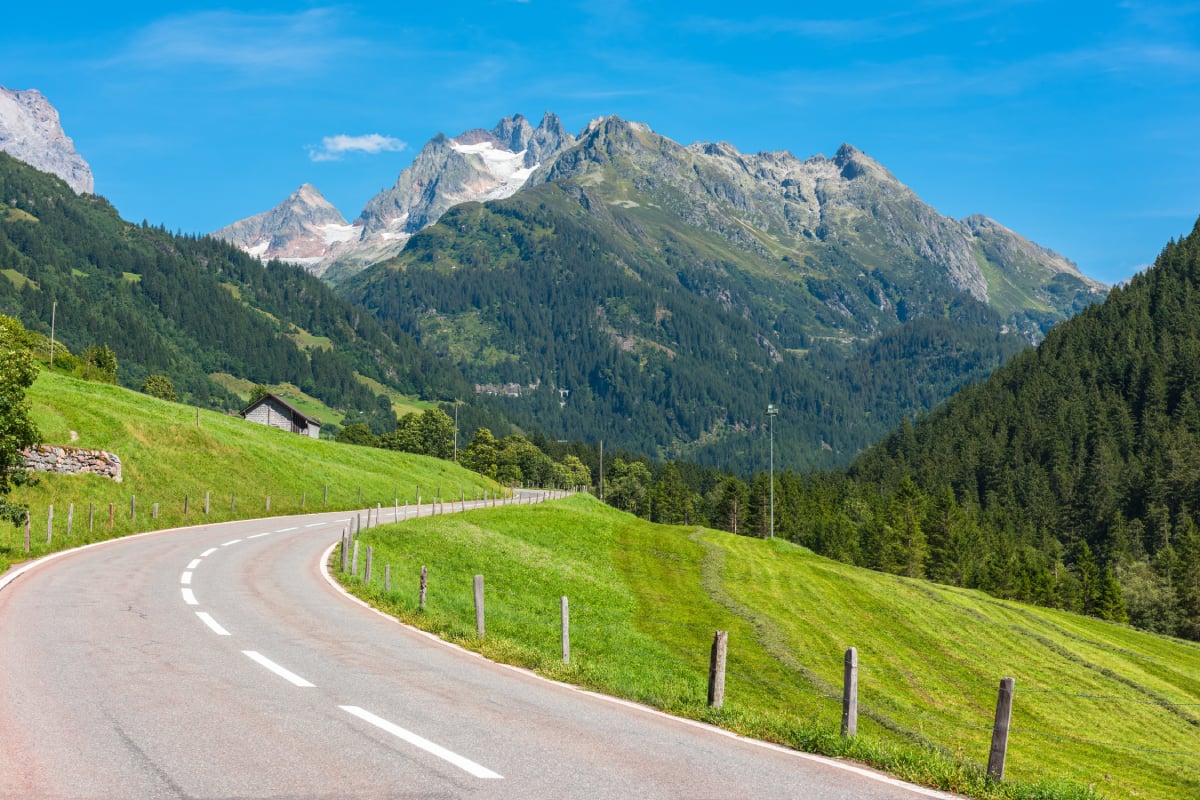 A winding country road curves through lush green hills toward a dramatic mountain range with rocky peaks and patches of snow. Dense forests cover the slopes beneath a clear blue sky.