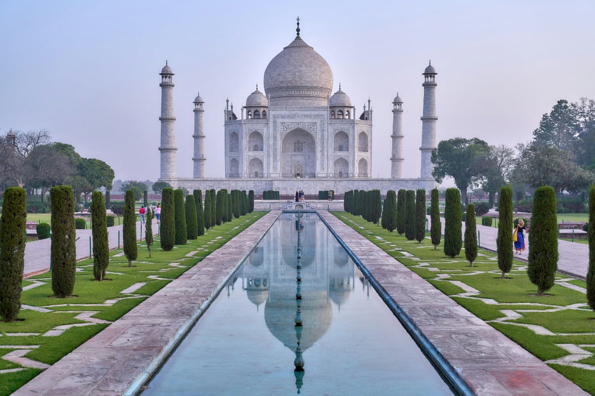 The Taj Mahal stands beyond a long reflecting pool and manicured gardens beneath a hazy morning sky in Agra, India.
