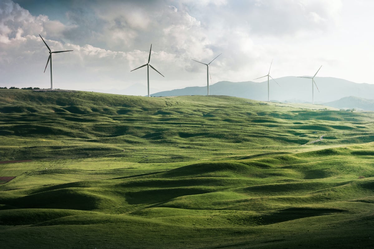 Wind turbines stand across rolling green hills beneath a cloudy sky with distant mountains on the horizon.