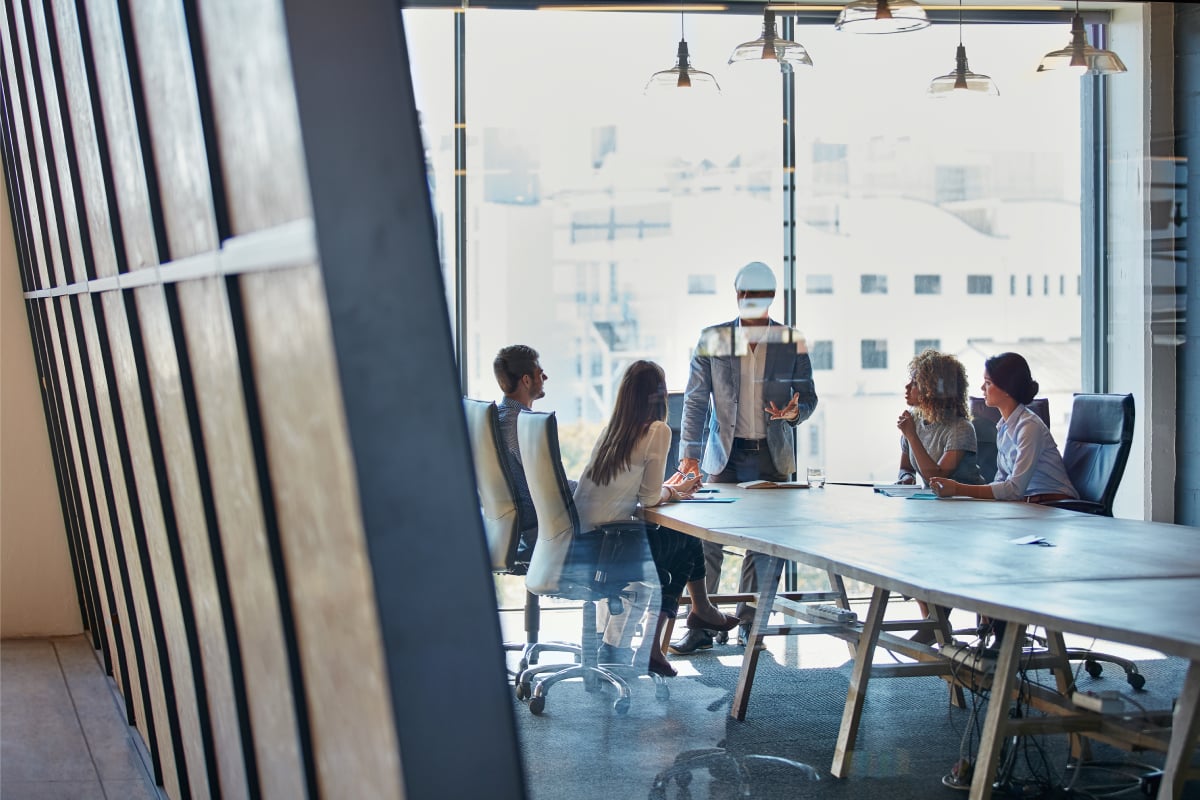 A group of five professionals sit around a conference table in a modern glass-walled office, listening to a colleague standing and speaking. Sunlight streams through large windows behind them, with a cityscape visible outside. The team appears engaged in a meeting or discussion, with notebooks and papers on the table.