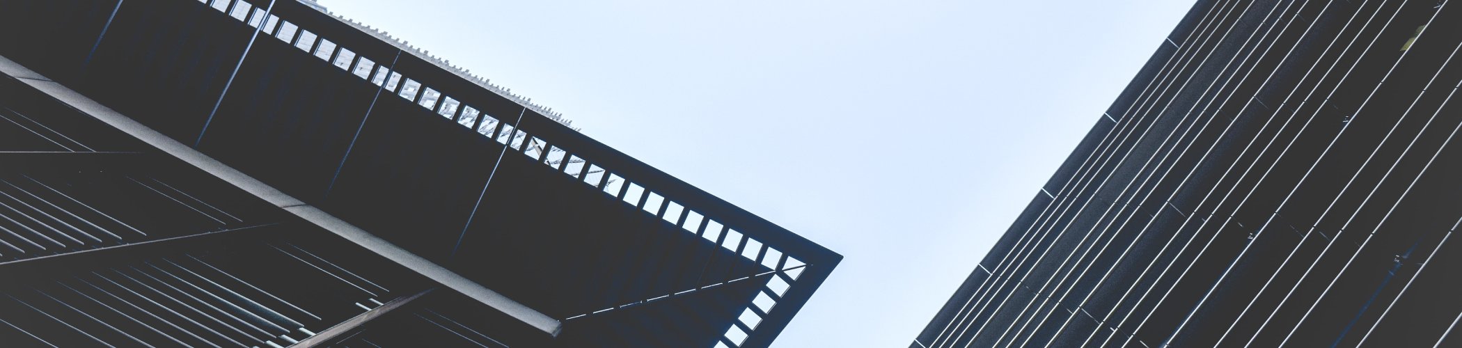 Upward view between modern buildings with geometric facades, creating intersecting lines against a bright sky.

