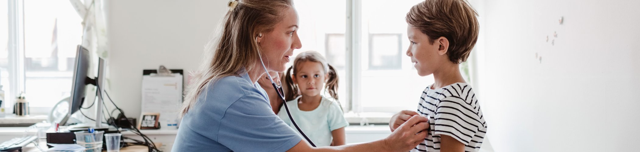 Healthcare professional using a stethoscope to examine a child, while another child stands in the background in a clinical room.

