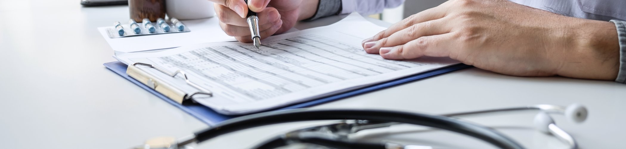 Healthcare professional writing on a medical record with a stethoscope on the table, illustrating consultation and patient care
