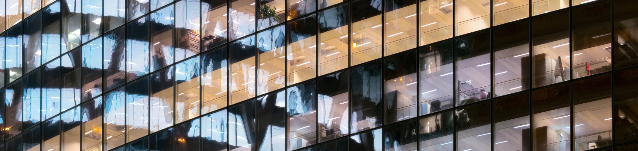 Glass facade of an office building with illuminated workspaces at night, illustrating corporate activity and financial markets
