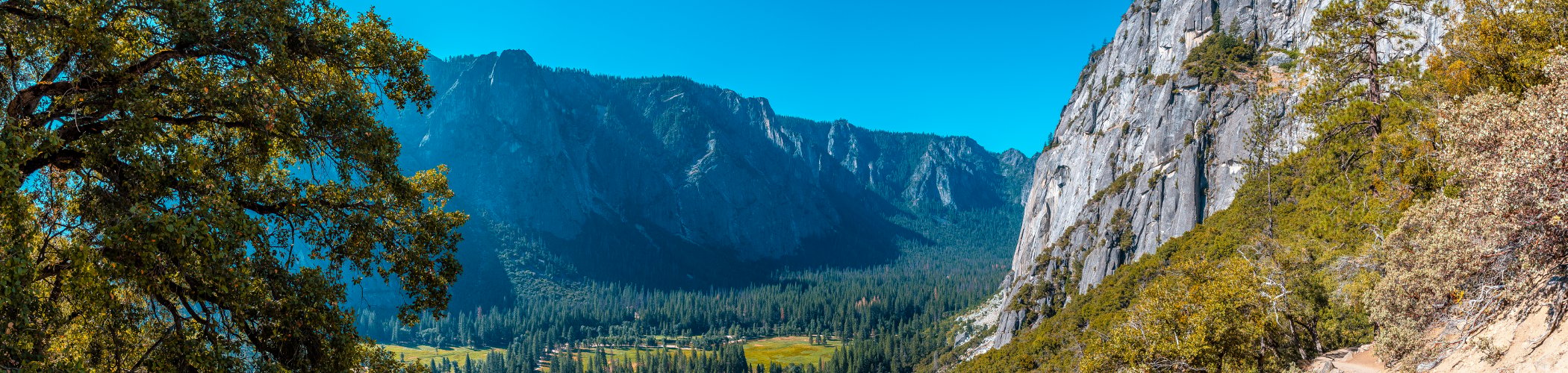 A scenic mountain valley viewed from a rocky hiking trail, with towering granite cliffs, dense evergreen forests, and a sunlit meadow below. The landscape is framed by trees under a clear blue sky.