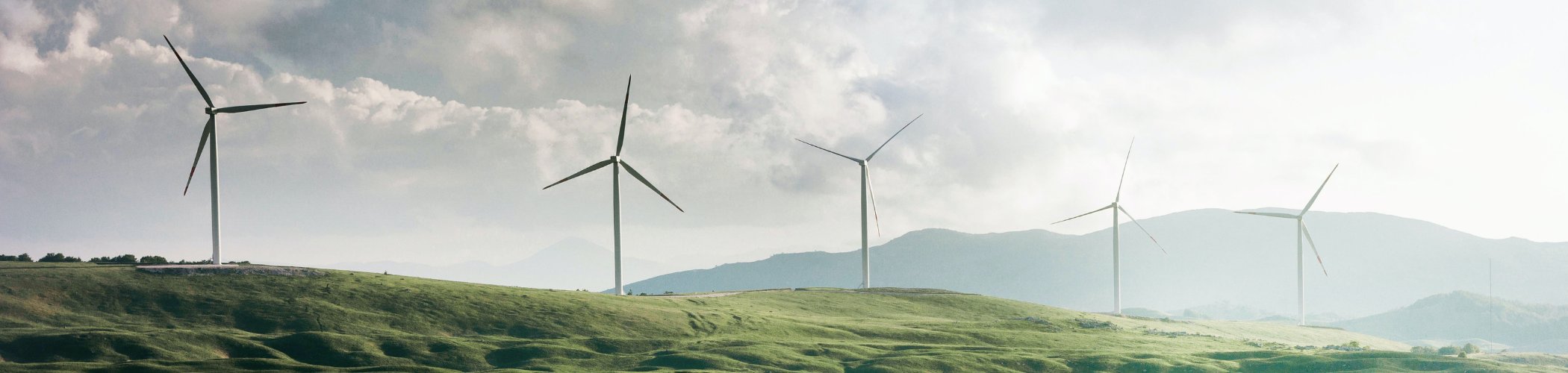 Wind turbines stand across rolling green hills beneath a cloudy sky with distant mountains on the horizon.