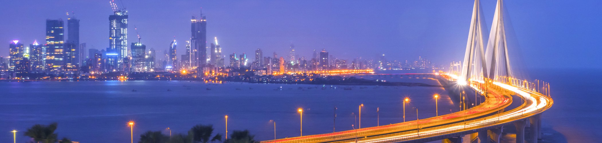 A cable-stayed bridge illuminated at night curves toward a city skyline across calm water beneath a cloudy blue sky.