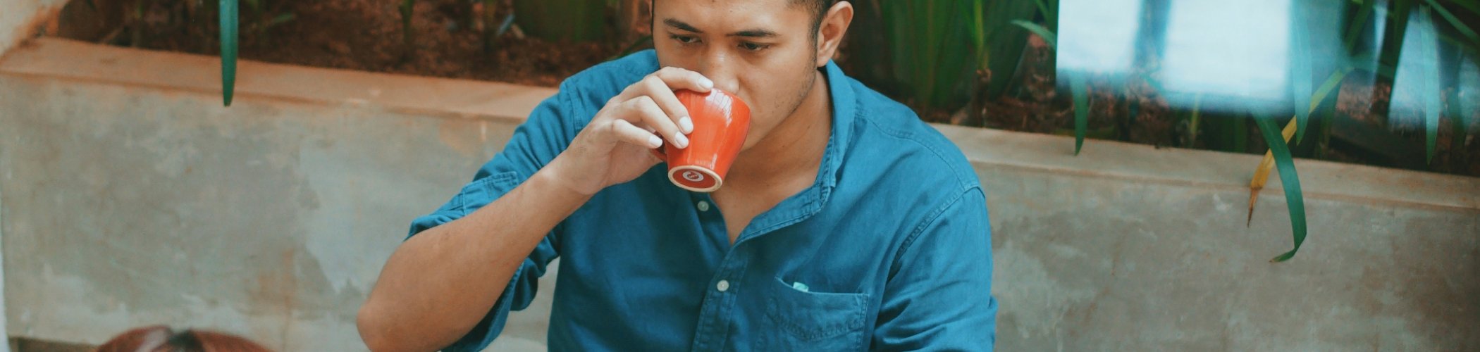Person working on a laptop while drinking coffee in an indoor space with plants, illustrating flexible work and coworking
