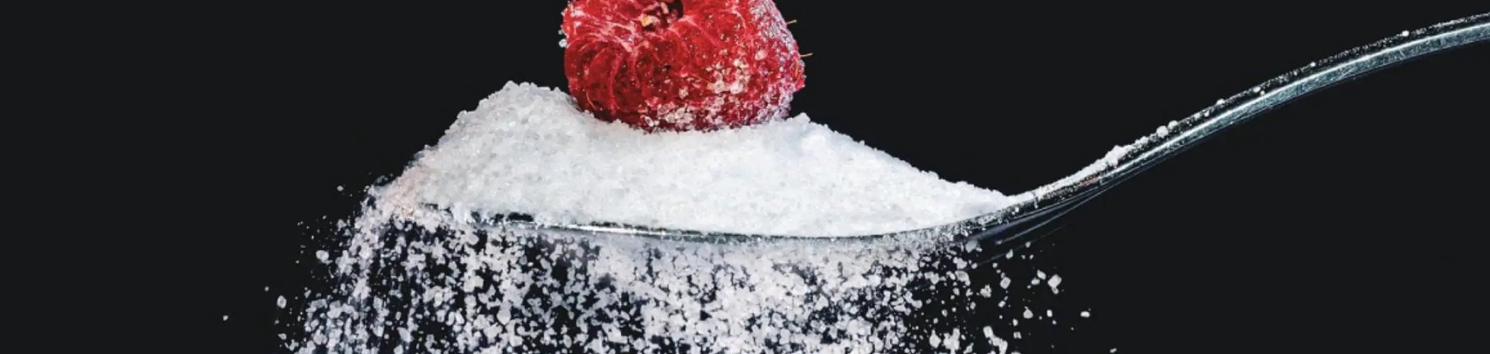 Close‑up of a spoon filled with granulated sugar and topped with a raspberry on a dark background