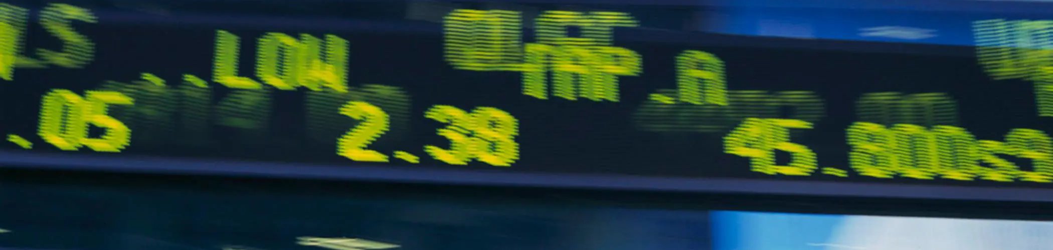 Close‑up of an electronic stock market display showing glowing green and yellow numbers, percentages, and abbreviations on a dark background.


