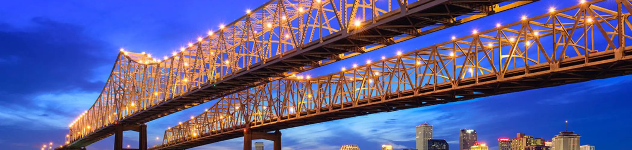 Illuminated bridge seen at dusk, stretching across a river with city buildings visible in the background.

